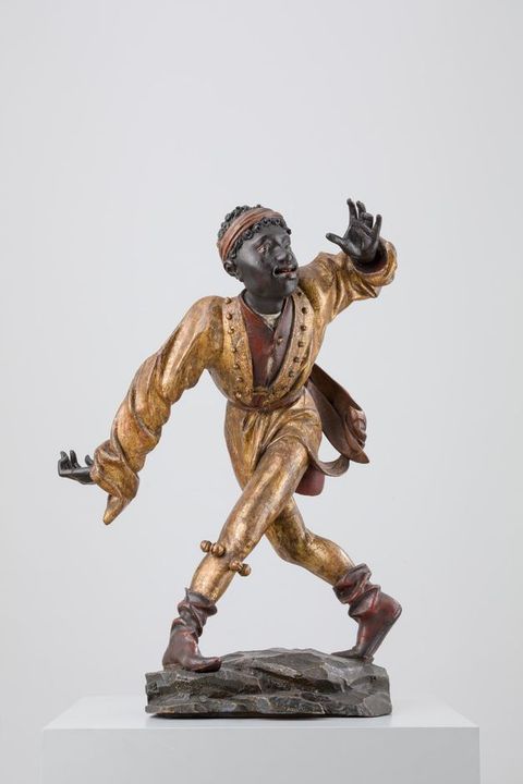 Sculpture. Carved from wood and then painted, a man with dark brown skin is shown mid-dance step. One leg crosses far in front of the other, feet planted wide. An extended arm matches the angle of the leg, and the other hand is raised. The person’s red lips are parted, and they look up and to our right.