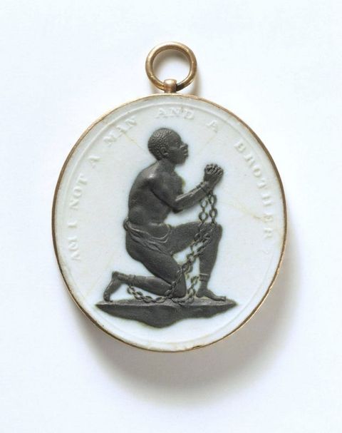 Medallion. In black against a white oval background, a man kneels and lifts his clasped hands. Chains connect shackles on his wrists to more on his ankles. His torso hunches forwards as he looks up. Raised lettering around the perimeter reads, “Am I not a Man and a Brother?” in all caps.