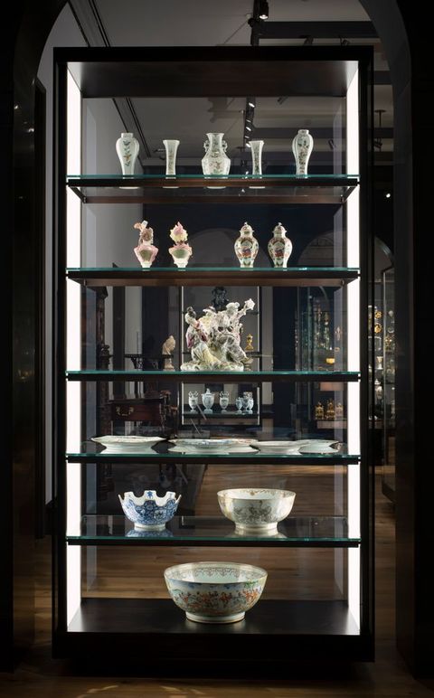 Installation. Six shelves hold porcelains in a case with glass on both long sides so we look through it. From the bottom up, the first two shelves hold bowls, and then a shelf has several plates. A sculptural piece is next, below two shelves of vases and urns.