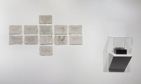 Installation. Two works are hung on a white wall. To our left, twelve rough-edged, rectangular pieces of paper appear nailed or tacked to the wall. A column of four papers down the middle is flanked by a square of four to each side. A black and possibly silver object is in a case to our right.