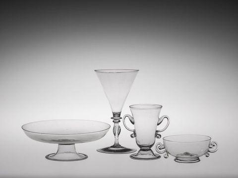 Glass. Two dishes and two glasses are clear blown glass. A tall posset glass and bowl have ear-shaped handles. The posset glass, a triangular wine glass, and a dish flare delicately at the lips.