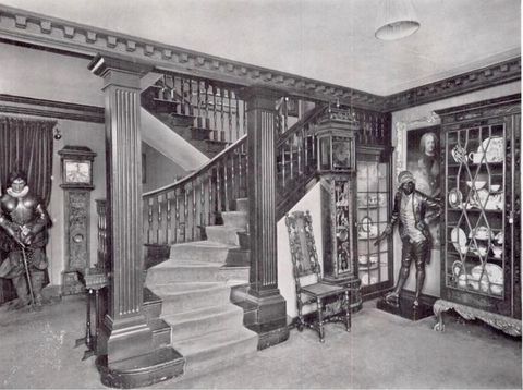 Black and white photograph. A zig-zagging staircase framed by fluted piers creates a corner, in which stand a chair, grandfather clock, two glass-fronted cabinets full of ceramic, and a life-sized, free-standing person. Details are difficult to make out in this image, but the person wears a coat with tails, knee-length breeches and stockings, and a long vest.